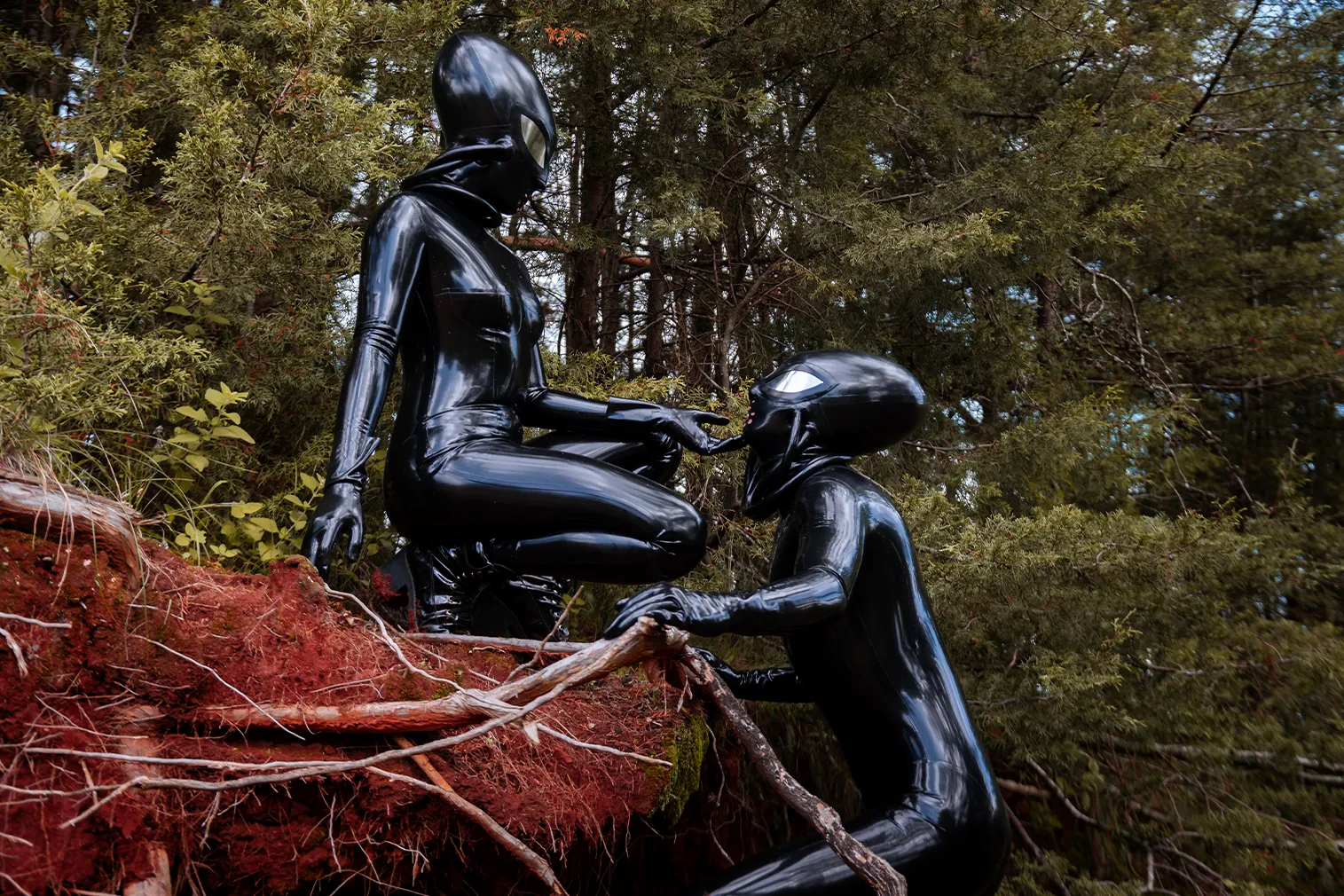 Dos personas con catsuit de látex negro y máscaras de alien, en un entorno de bosque